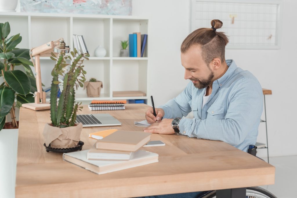 happy disabled man on wheelchair writing in notebook at workplace